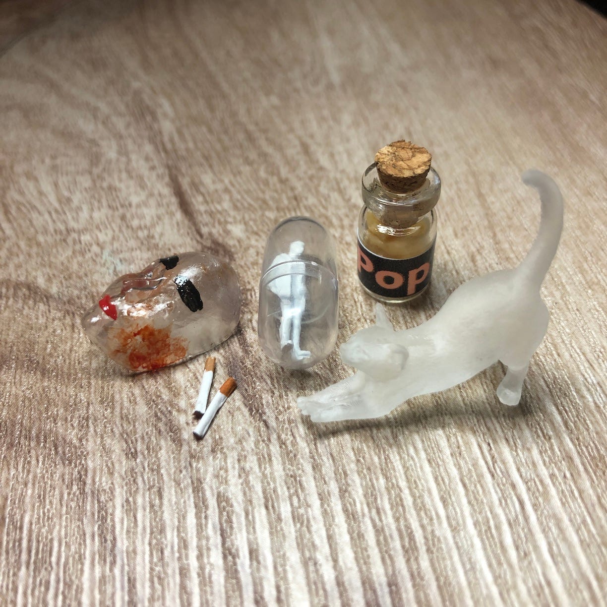 Miniature clear, bloody mask, ghost cat, cigarettes, person in a capsule, and jar of "pop" (which looks like peeled Caucasion skin) on a gray wooden table