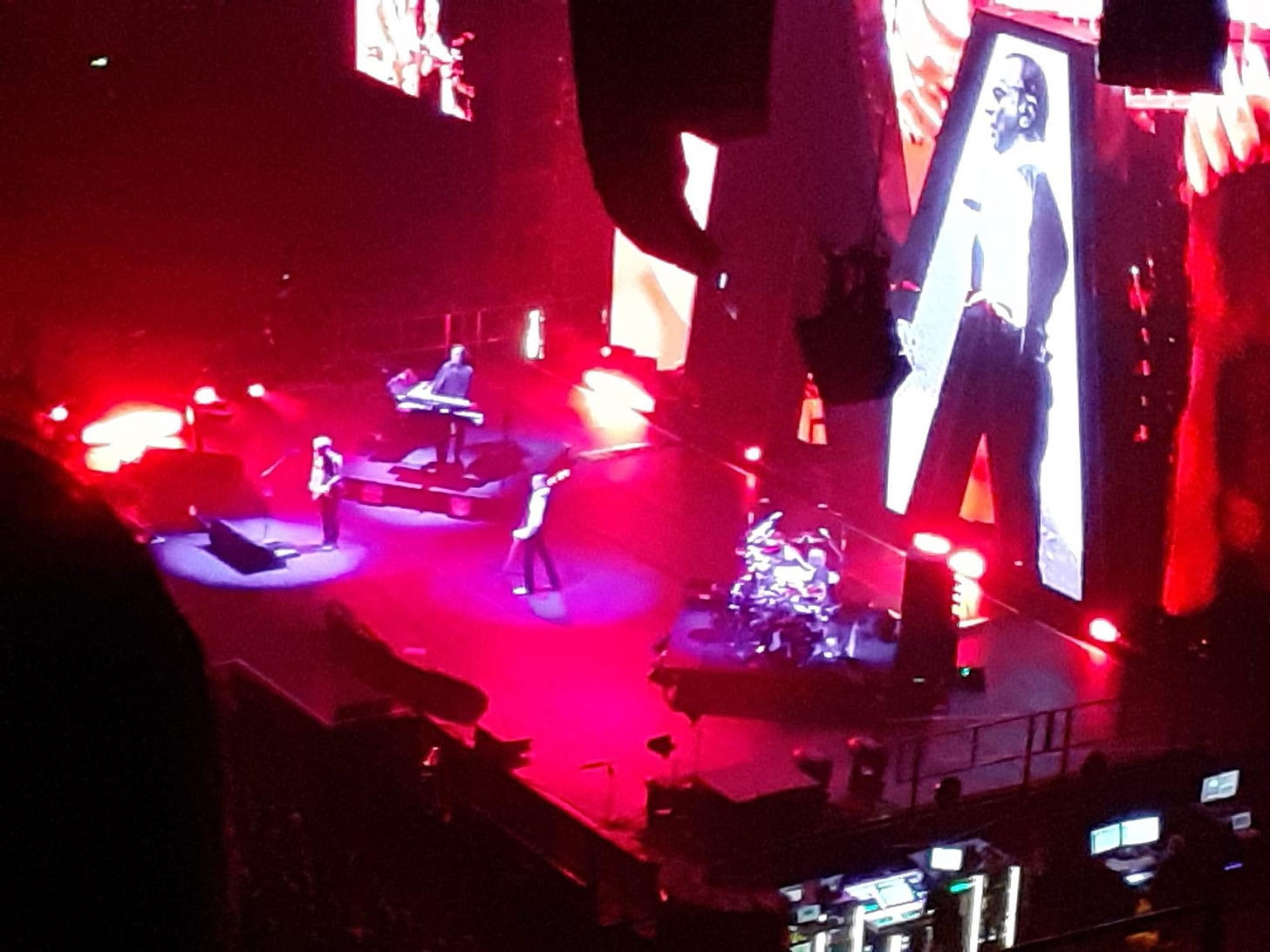 A distant view of the band depeche mode on a red lit stage, one person playing a guitar, one the keyboards, one the drums, and a singer. There is a large screen in the background with a bit M on and a video projection of the lead singer
