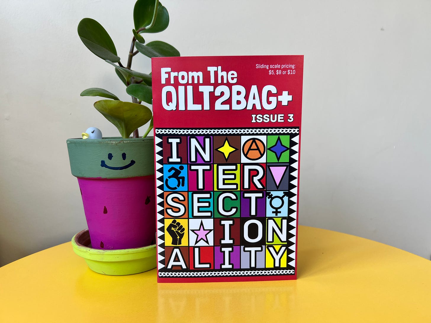 From The QILT2BAG+ issue 3, featuring cover art by Aaron Beaumont.  The 'zine is standing up on a yellow table with a thick leaved green plant in a pink and green painted lant pot behind it. 