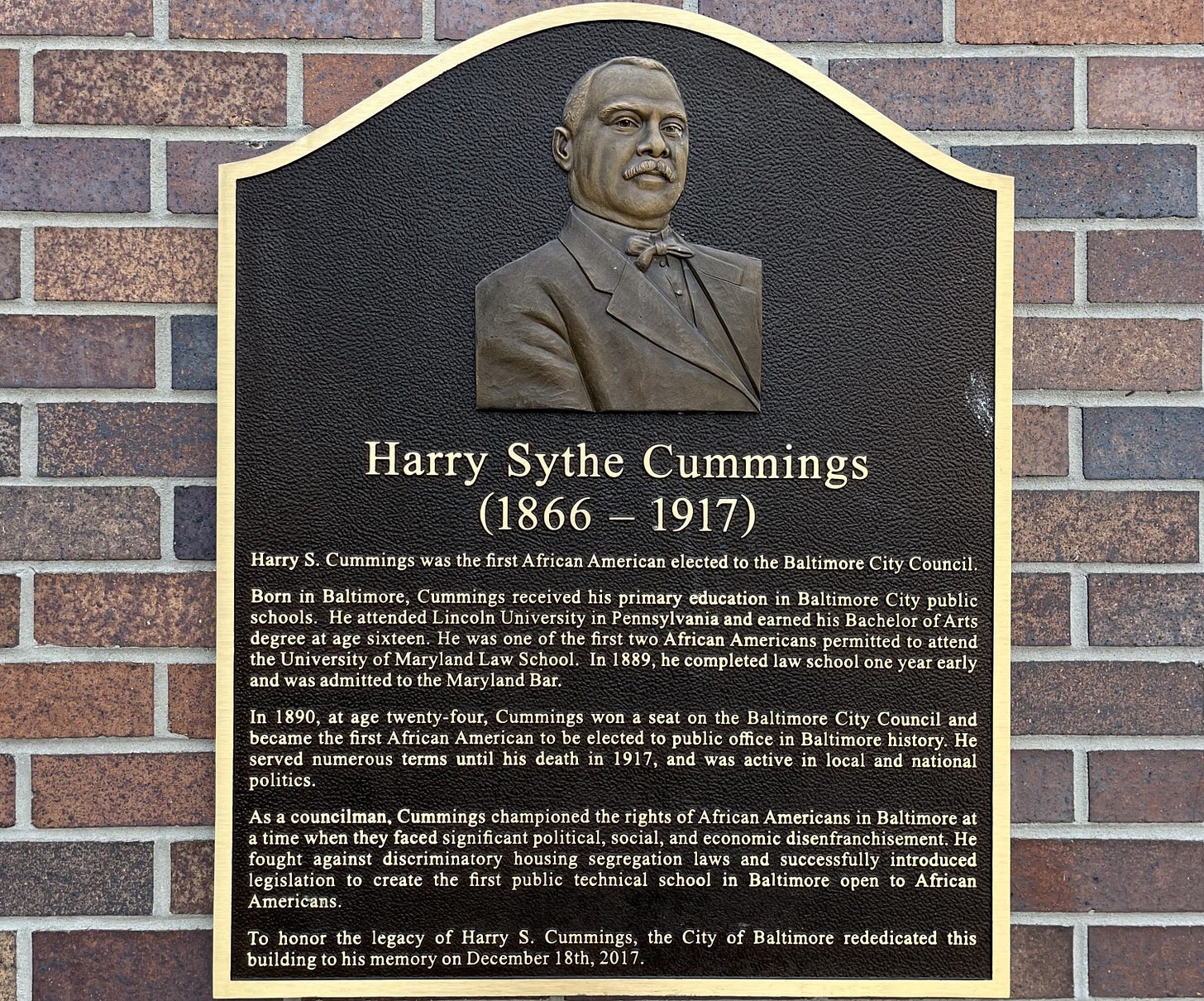 Photo of a historical marker on the side of a brick building titled: “Harry Sythe Cummings (1866-1917).” It includes an engraved image of Cummings near the top of the marker.