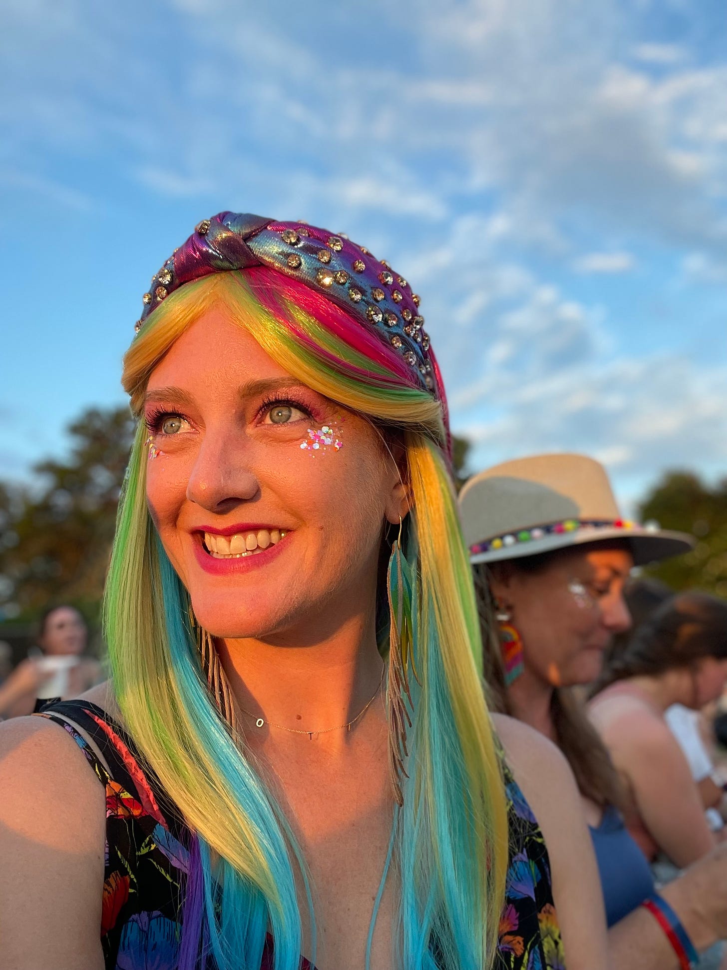 Becky wears a long, silky rainbow wig and a shimmering, rhinestoned headband. She beams into the distance, chunky glittery shimmering below her green eyes. Soft clouds float across the blue sky behind her and a friend in a fun hat is over her shoulder.