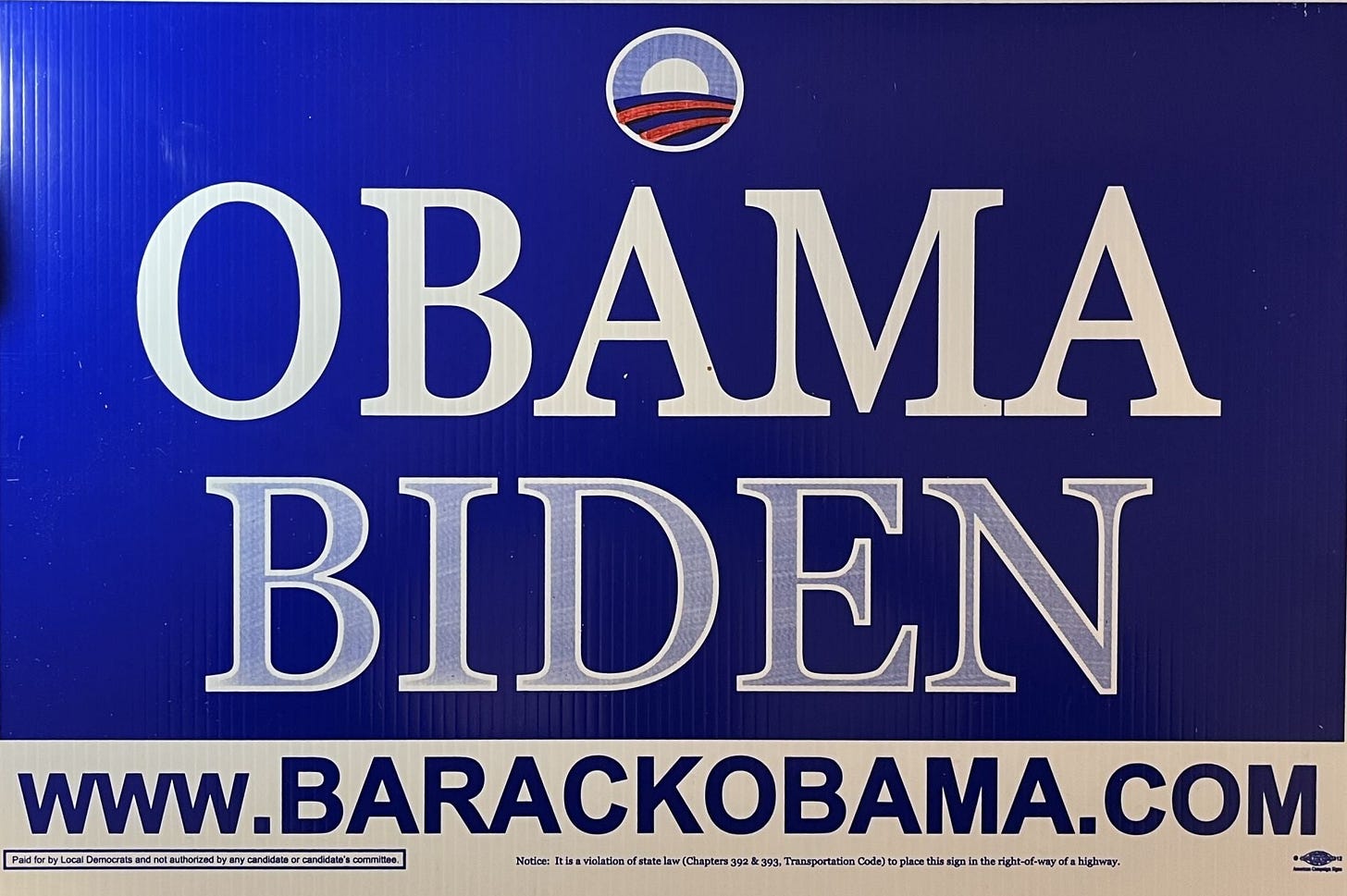 This Obama-Biden campaign sign has the standard, circular, red-white-and-blue sunrise-over-America icon at the top, a navy-blue background, Obama's name (in white type) below it, Biden's surname (in white type) below that, and the website URL www.BARACKOBAMA.COM (in blue type over a white background) at the bottom. This Obama-Biden campaign sign has the standard, circular, red-white-and-blue sunrise-over-America icon at the top, a navy-blue background, Obama's name (in white type) below it, Biden's surname (in white type) below that, and the website URL www.BARACKOBAMA.COM (in blue type over a white background) at the bottom.