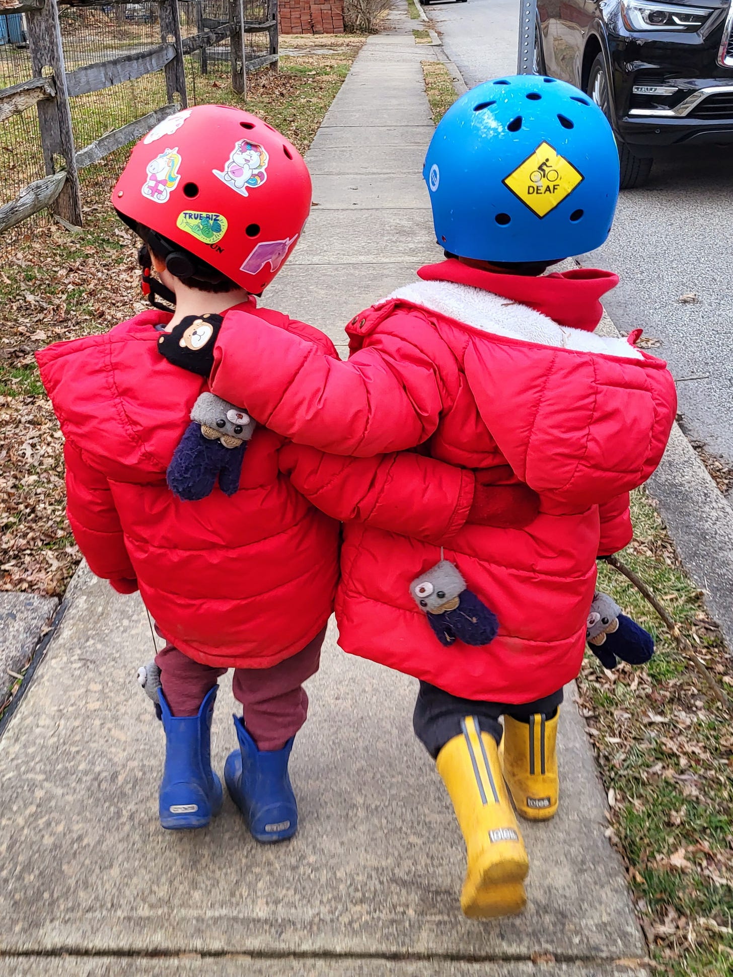 ID: Two brothers in rainboots, matching red coats, and bike helmets walk with their arms around each other. One has a sticker on his helmet that says “Deaf.” 