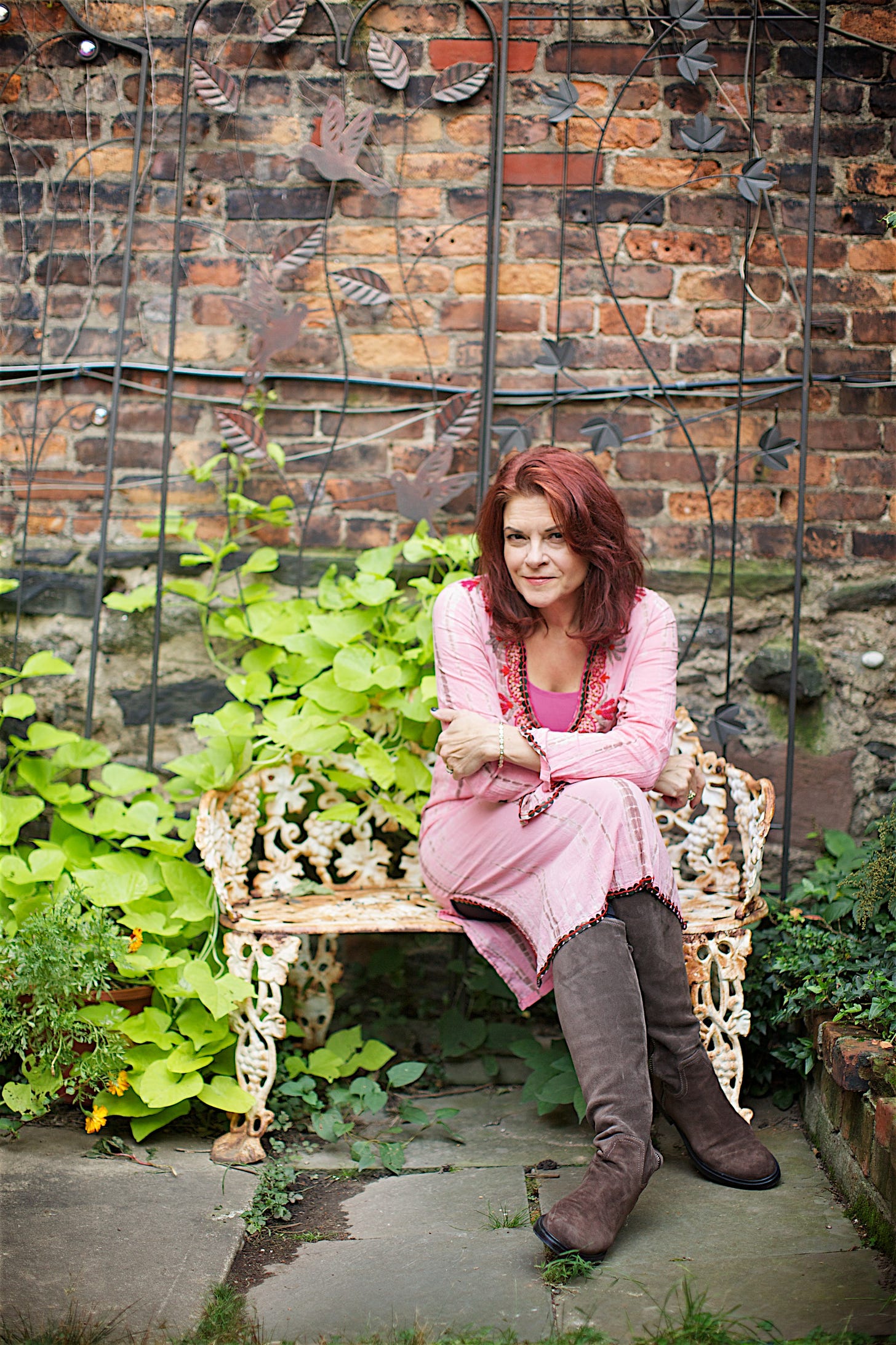 Rosanne Cash sits on a bench in her garder in a pink outfit and grey boots