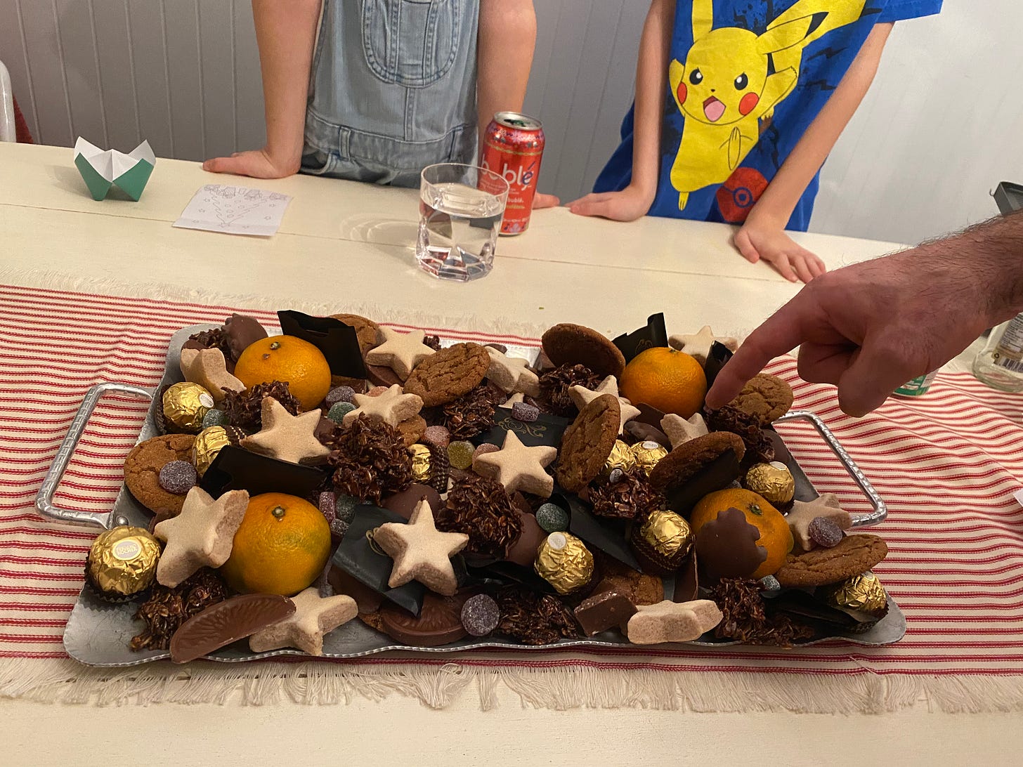 On a table laid with a white tablecloth and a red and white table runner, a metal tray with artfully arranged treats on it: zimtsterne, haystacks, ginger cookies, chocolates, oranges, small round gummies. Adrian's hand is pointing things out in the right side of the frame, and two kids stand in the background, waiting impatiently.