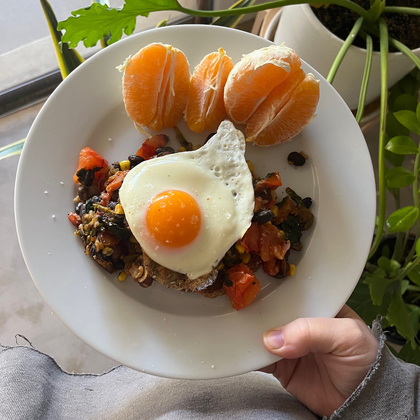 Me holding a white plate with a sunny side up egg over beans, corn, tomatoes, mushrooms with an orange on the side.