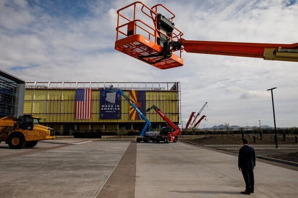 Construction machinery and a lone figure standing on pavement in front of an industrial building. A “Made in America” banner hangs between a U.S. flag and an Arizona state flags down the side of the building.