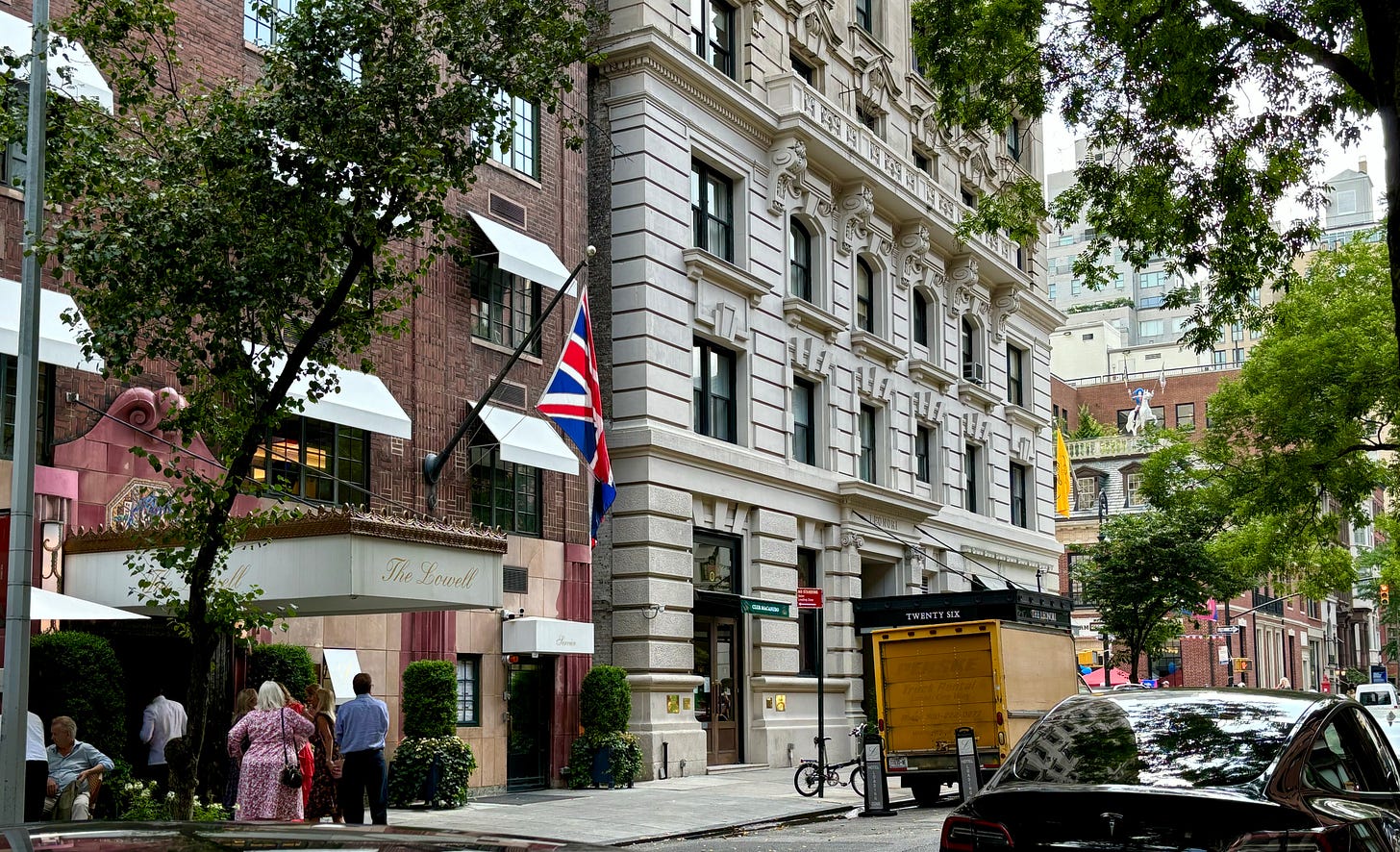 A stately-looking building. There is a white awning with The Lowell written in gold script. Above it hangs the British flag. Beyond the fine white building on the corner is a barely visible street fair, as well as a statue of Napoleon on a white horse on the roof of the Hermes on 63rd street across Madison Avenue.