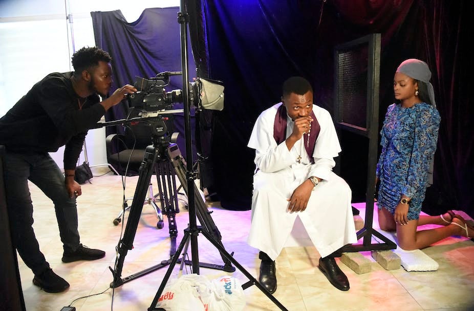 Man peers through a camera shooting a man sitting on a chair and a woman on her knees.