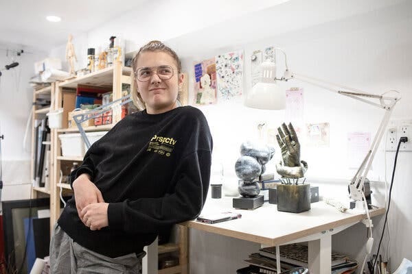 Aidan, wearing a black sweater, is leaning on a white art table, on which are several small metal sculptures. Paper works hang on the white walls and in the background, a tall wooden case holds various art supplies.