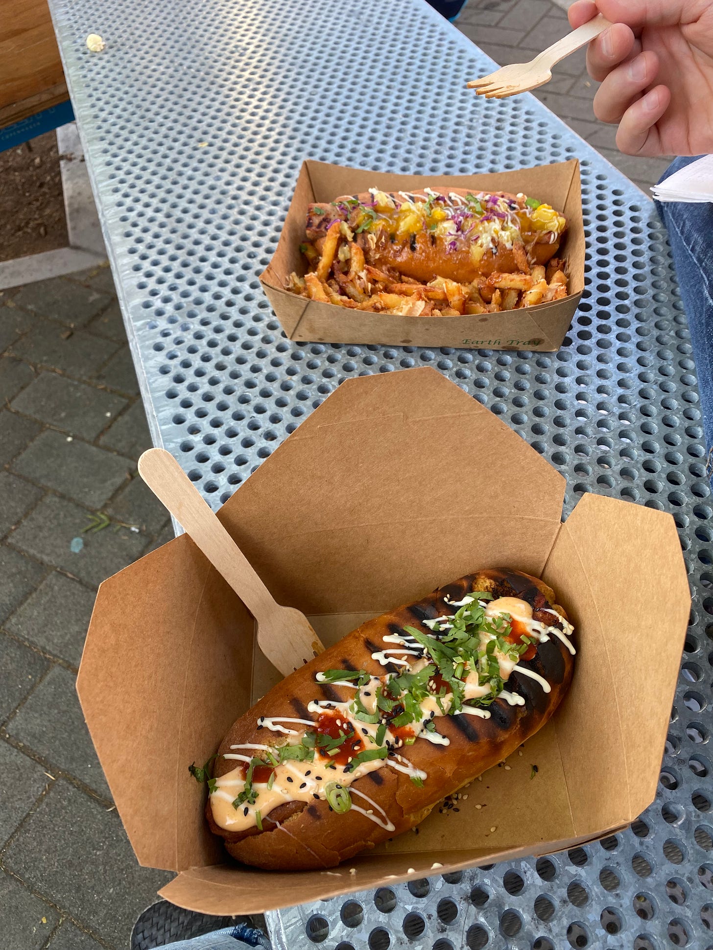 On a metal bench, two paper takeout containers: in the front, a hero with drizzles of spicy mayo, regular mayo, and hot sauce, dusted with green onions and sesame seeds; in behind it, a hero with mango sauce and green onions, and fries drizzled with mayo and sprinkled with nori. Jeff's hand holds a wooden fork over the container.