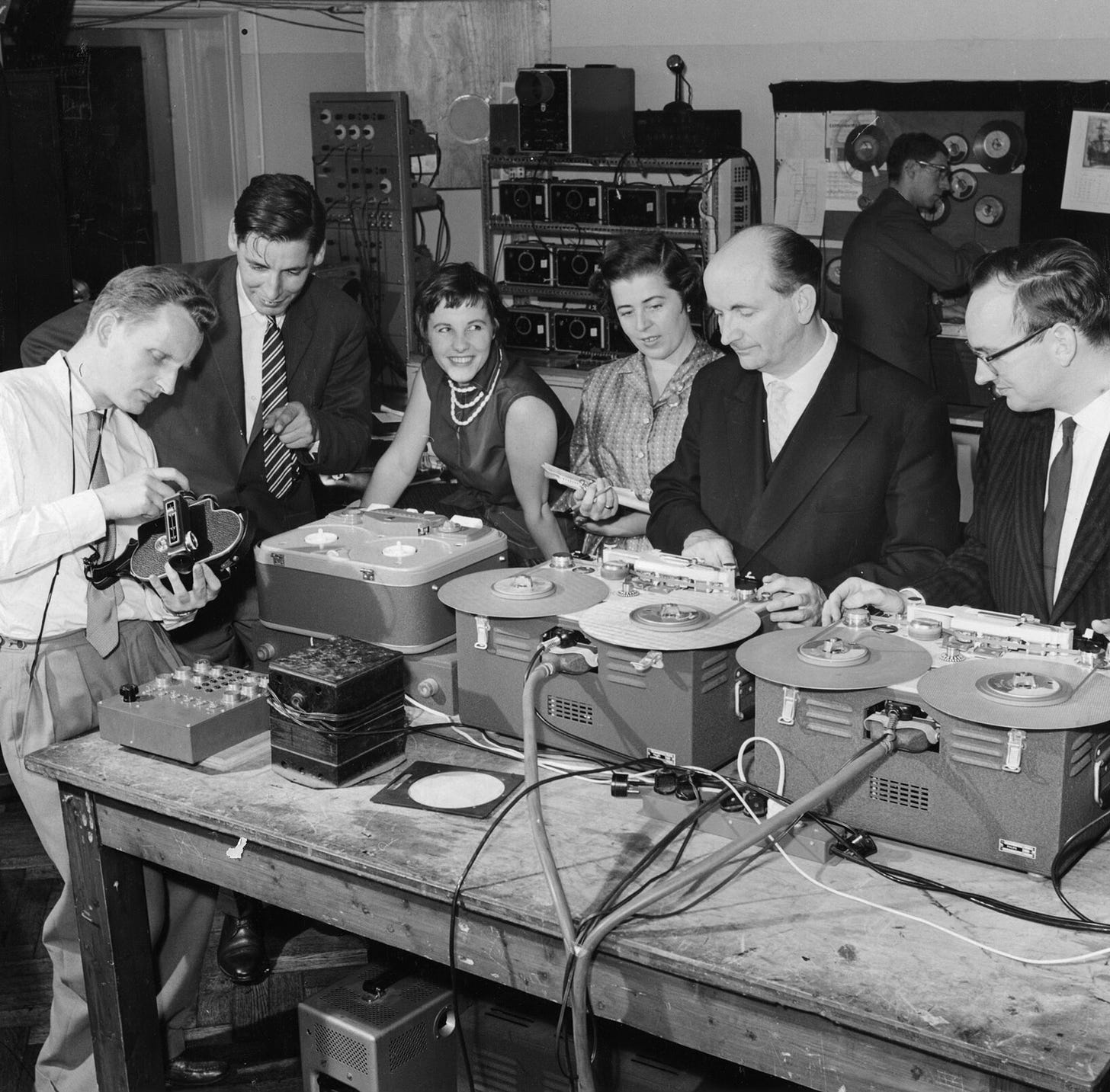 Deze zwart-wit foto toont een groep mensen in een studio- of laboratoriumomgeving, verzameld rond verschillende bandrecorders met spoelen. De personen zijn formeel of semi-formeel gekleed, typisch voor het midden van de 20e eeuw. De centrale figuren zijn bezig met het bedienen van de bandmachines, mogelijk met het opnemen of bewerken van audio, terwijl een andere persoon links apparatuur inspecteert of aanpast, waarbij hij een filmcamera of ander soort apparaat vasthoudt. Op de achtergrond zijn meer elektronische apparaten en kabels zichtbaar, wat bijdraagt aan de technische sfeer van de scène. De omgeving lijkt een professionele of experimentele audioproductiestudio te zijn.