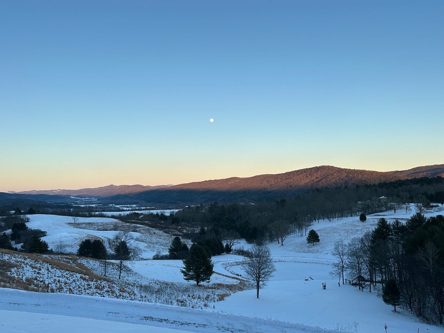 an image of a darkening blue sky and a full white moon rising above the darkened mountains, with snow in the foreground 