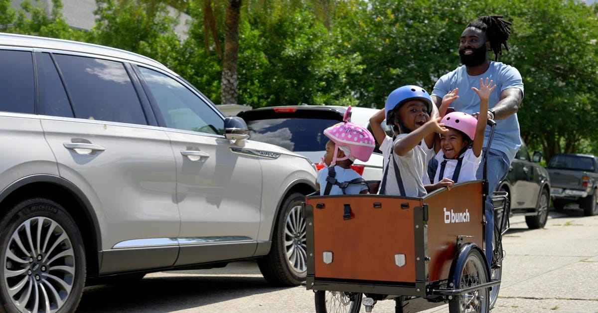 A Black parent riding a cargo bike containing three Black children wearing helmets, with arms raised and joyful expressions.