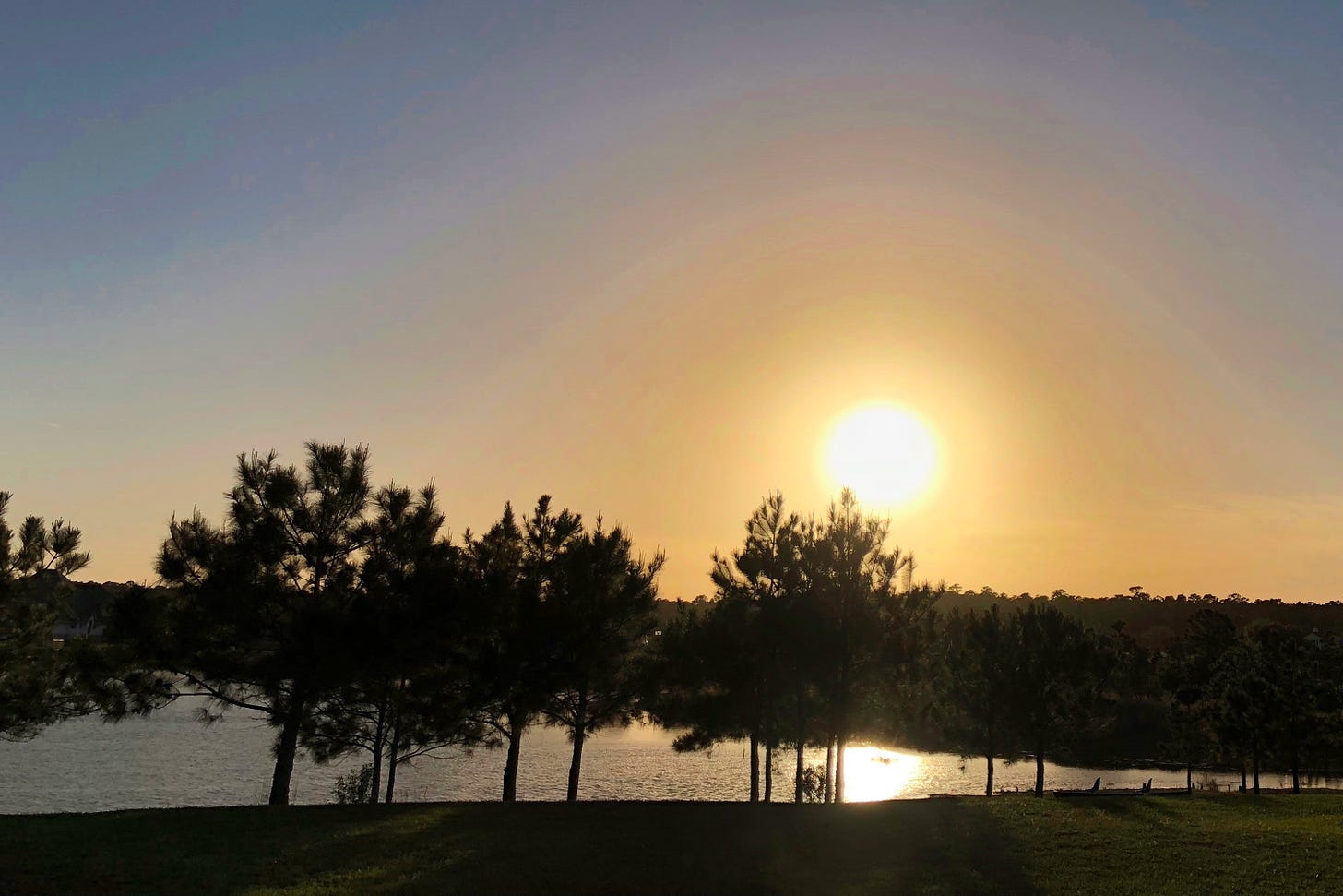 A sunset with the sun just above the horizon line casting an orangish/yellow halo across the blue sky and the sun’s reflection in the lake in the foreground