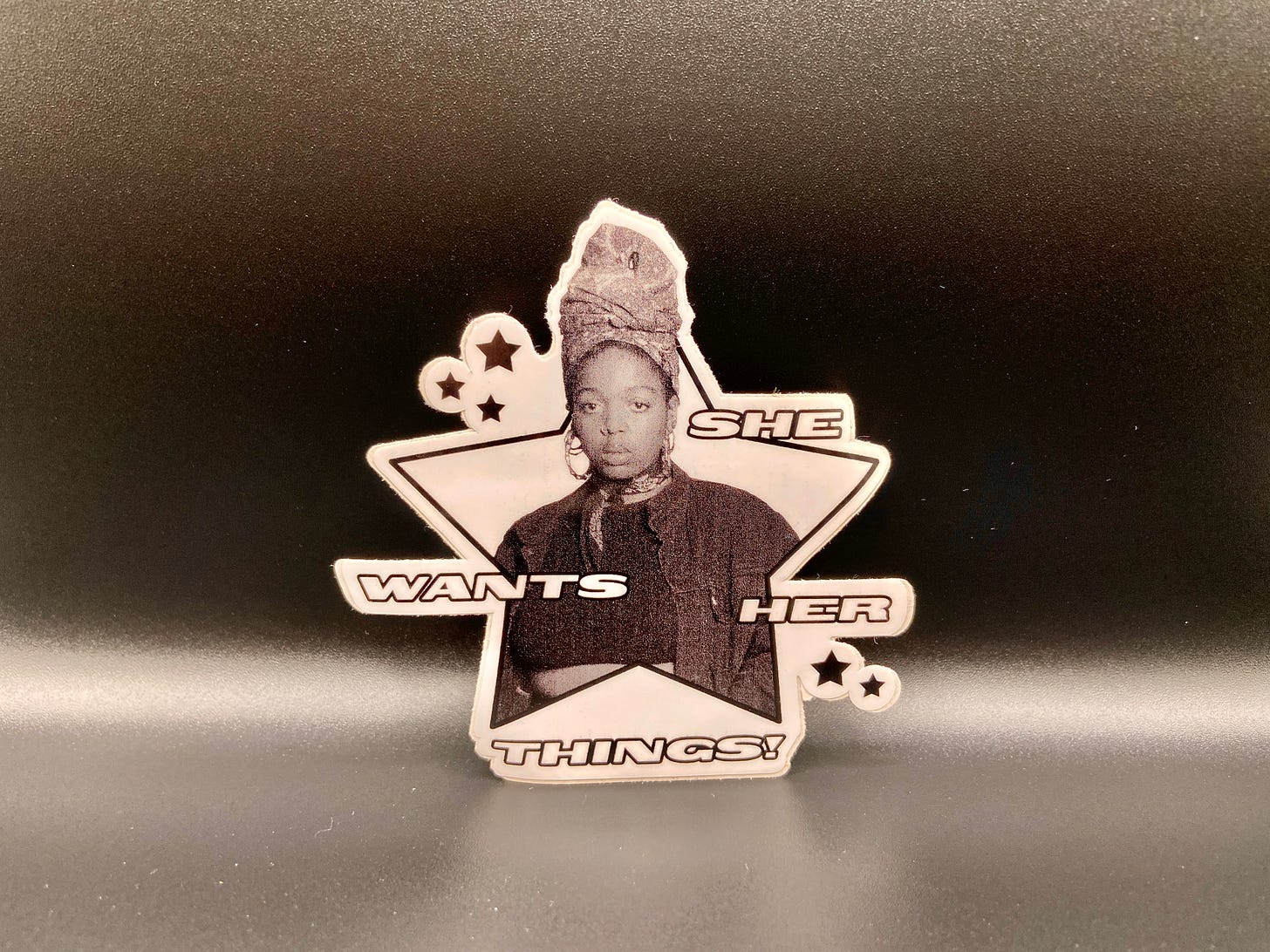 a star-shaped sticker with a Black woman wearing a turban on her head, surrounding by the text “SHE WANTS HER THINGS.”