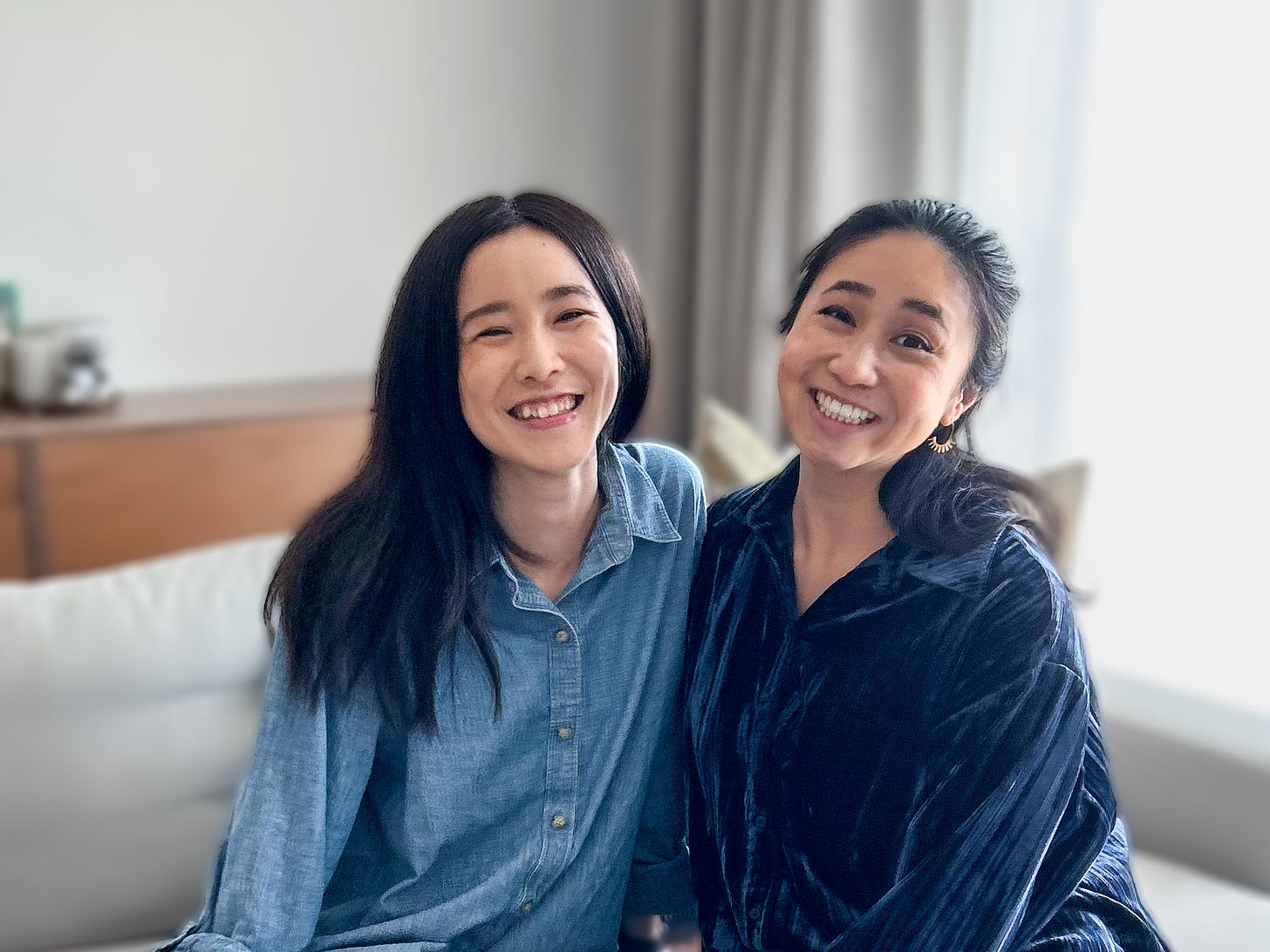 Two woman both with black hair sitting on a couch smiling at the camera