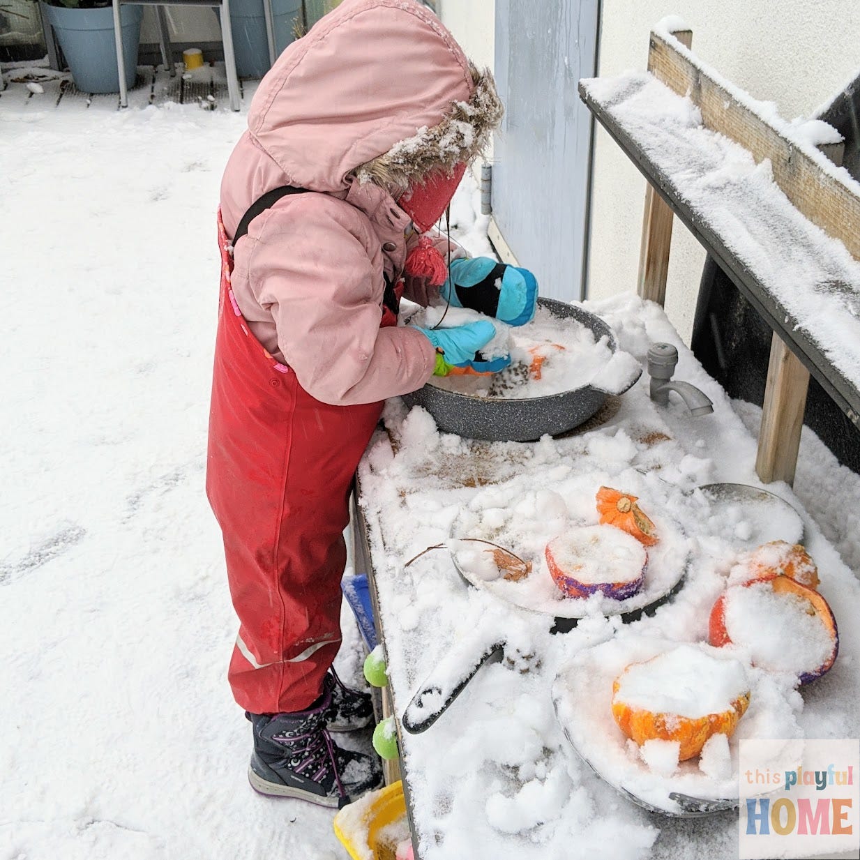 a young girl in a pink coat and red overalls plays in a snow covered mud kitchen