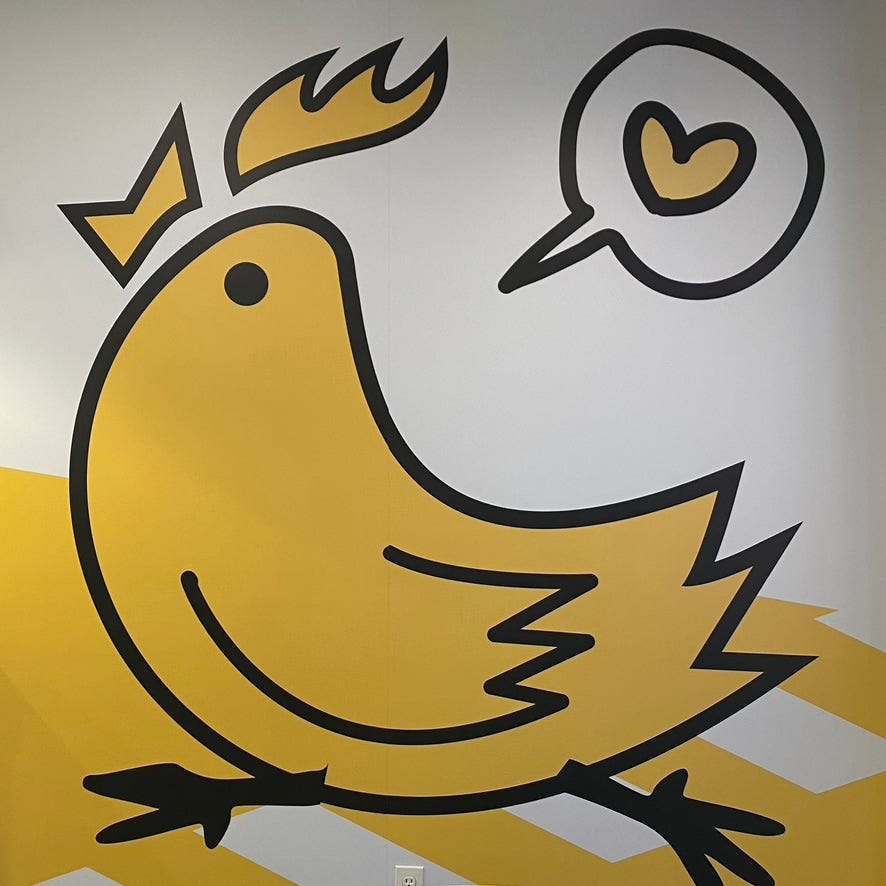 A cartoon chicken in bright yellow and bold black outline. It appears to be running somewhere in great excitement. Over its head is a speech bubble with a heart inside it.