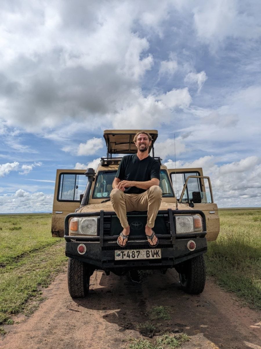 a smiling young man with a beard and flip-flops, sitting on the front of a safari jeep on an open plain with puffy clouds in the sky