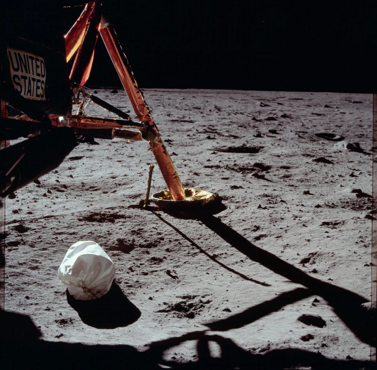 The cratered grey surace of the moon against the deep black of endless space. In the foreground are the reddish-legs of the moon lander module and a white bag of trash lying haphazardly against the moon's surface.