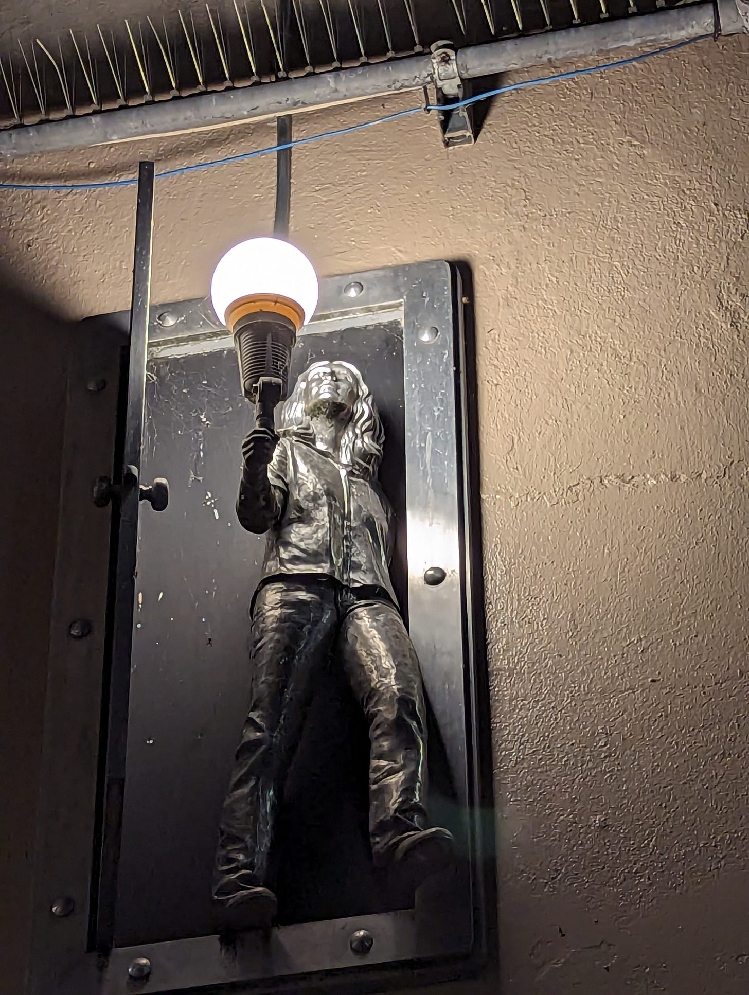 A lamp set high in a wall that is designed to look like a pewter woman with long hair who is holding a lamp in one hand as she pushed open a door and looks out into the world.