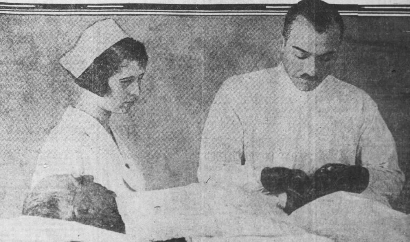 A photograph showing an elderly man lying on a bed with white linen. A male surgeon with white garments and dark surgical gloves is performing a procedure on the man's abdomen. To on the left of the image is a woman in nurse's uniform, including a traditional folded cap. She is looking at the region of the procedure.