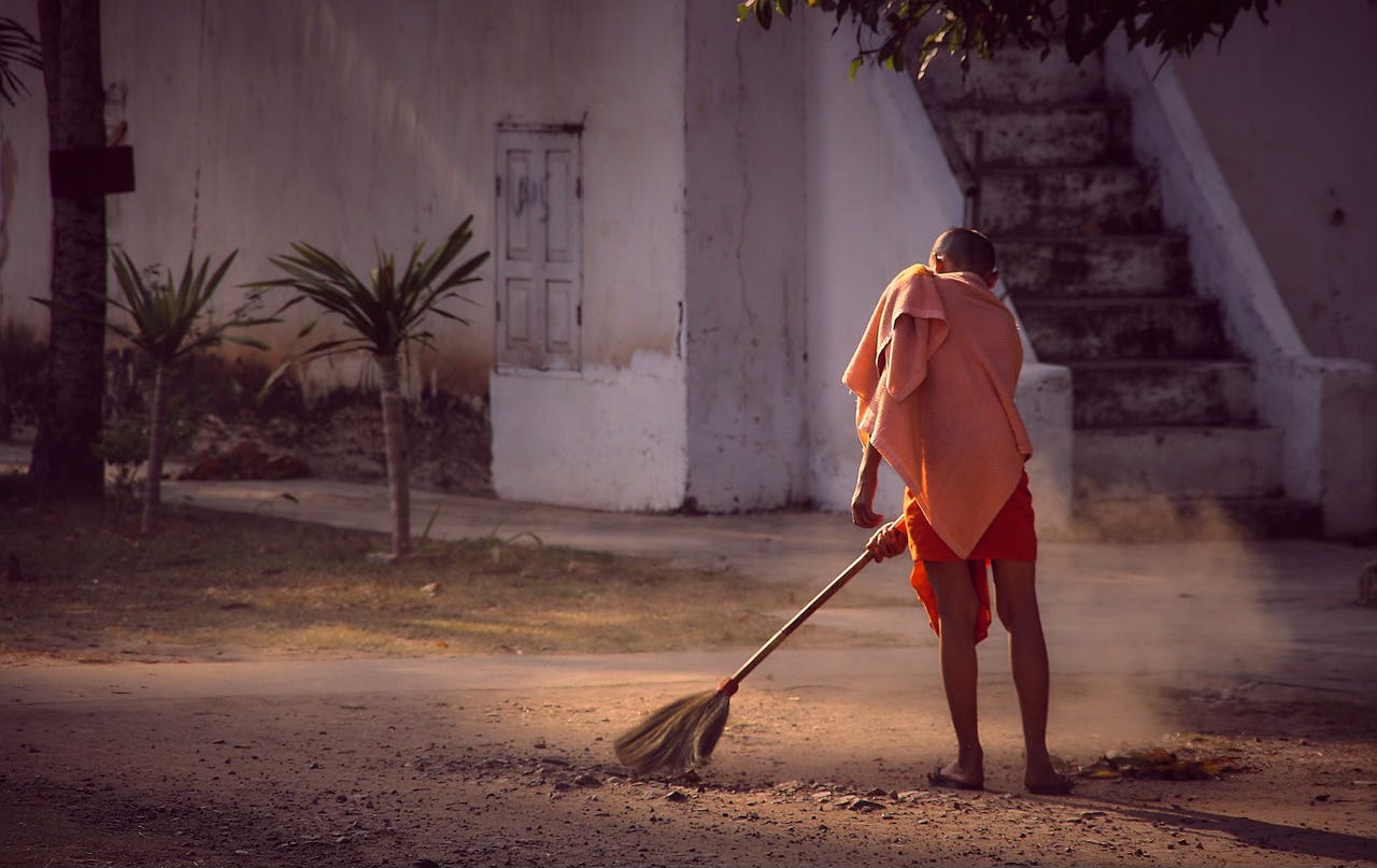 A young man in orange monk's robes sweeps the dirt in front of a whitewashed monastery with palm trees in the background.