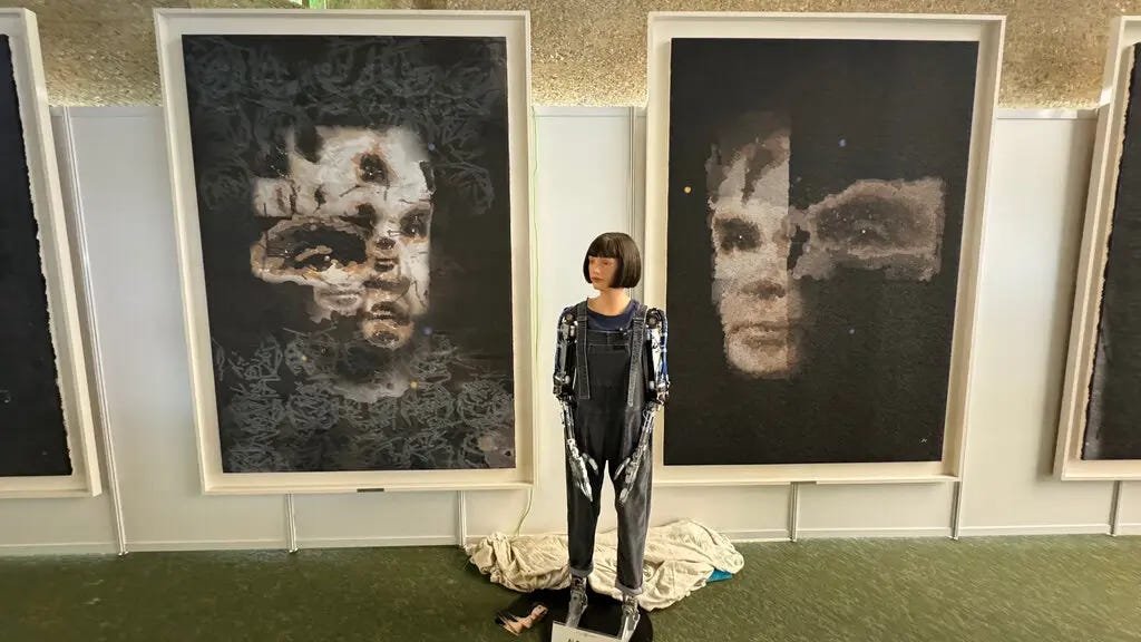 A humanoid robot wearing overalls and a bob haircut wig stands in front of two large semiabstract portraits of Alan Turing.