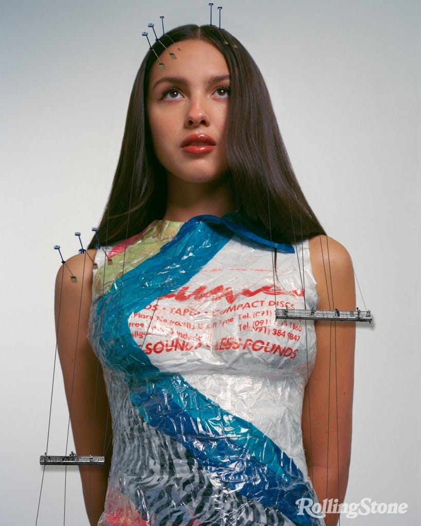 In just one weird portrait from Rolling Stone, Olivia Rodrigo stands stiffly, looking slightly up, dressed in… what appears to be trash (?) while two scaffolds of black and white window washers (??) hang badly Photoshopped from her shoulders. I am genuinely baffled at what’s supposed to be going on here.
