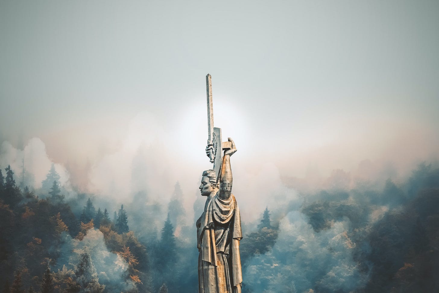 Fotografia em cores do Monumento à mãe pátria em Kiev, na Ucrânia. A estátua de um homem de braços erguidos, segurando um escudo e uma espada, está cercada de um nevoeiro. Ao fundo, árvores. Fotografia em cores do Monumento à mãe pátria em Kiev, na Ucrânia. A estátua de um homem de braços erguidos, segurando um escudo e uma espada, está cercada de um nevoeiro. Ao fundo, árvores.