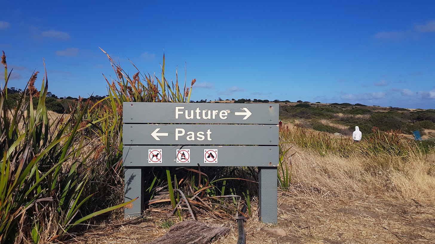 Image description: a gray sign outdoors that has Future with an arrow to the right and past with an arrow to the left. It reminds me of beach signage.
