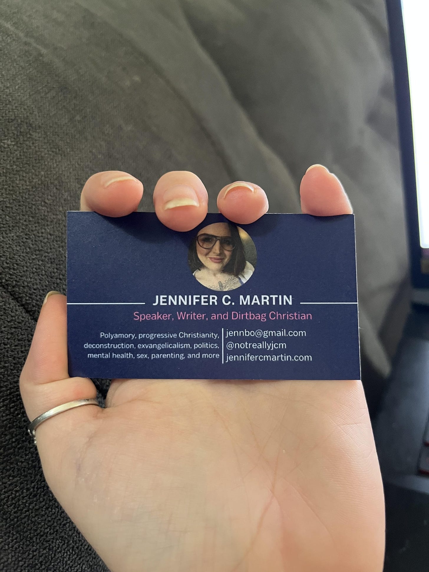 a business card (mine) in a woman's hand against a grey couch background. there's a photo of jennifer c. martin, with the word Jennifer C. Martin underneath, speaker, writer, and dirtbag christian, contact info and website info, and the words "polyamory, progressive Christianity, deconstruction, evangelicalism, politics, mental health, sex, parenting, and more"