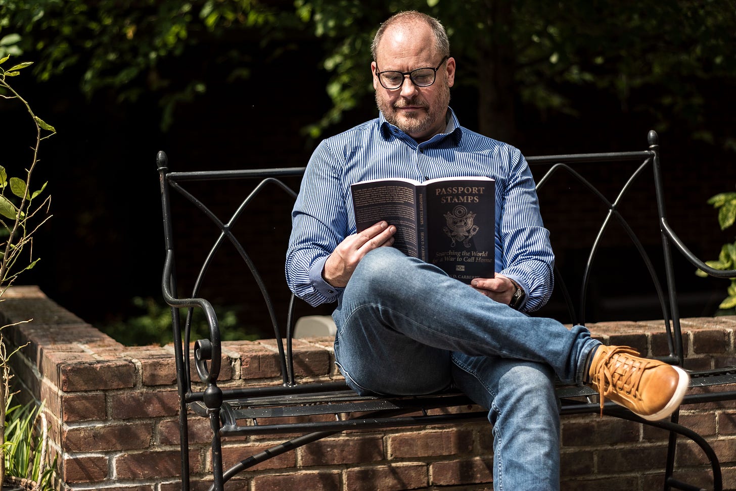 Photo of Sean Carberry sitting on a metal bench by a small brick wall and trees reading a copy of the book Passport Stamps