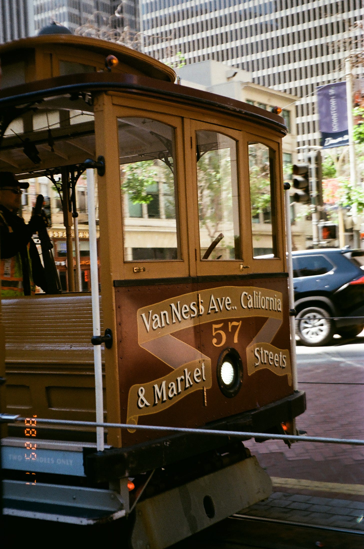 A close up film photo of the San Francisco's Van Ness Ave. California and Market Street  brown cable car. The conductor is in the cable car holding the lever, wearing a blue cap and a neon yellow with orange accent vest. The cable car is in the foreground of urban buildings. A small date watermark is on the left bottom corner with 88, 29, and 1 printed vertically. 