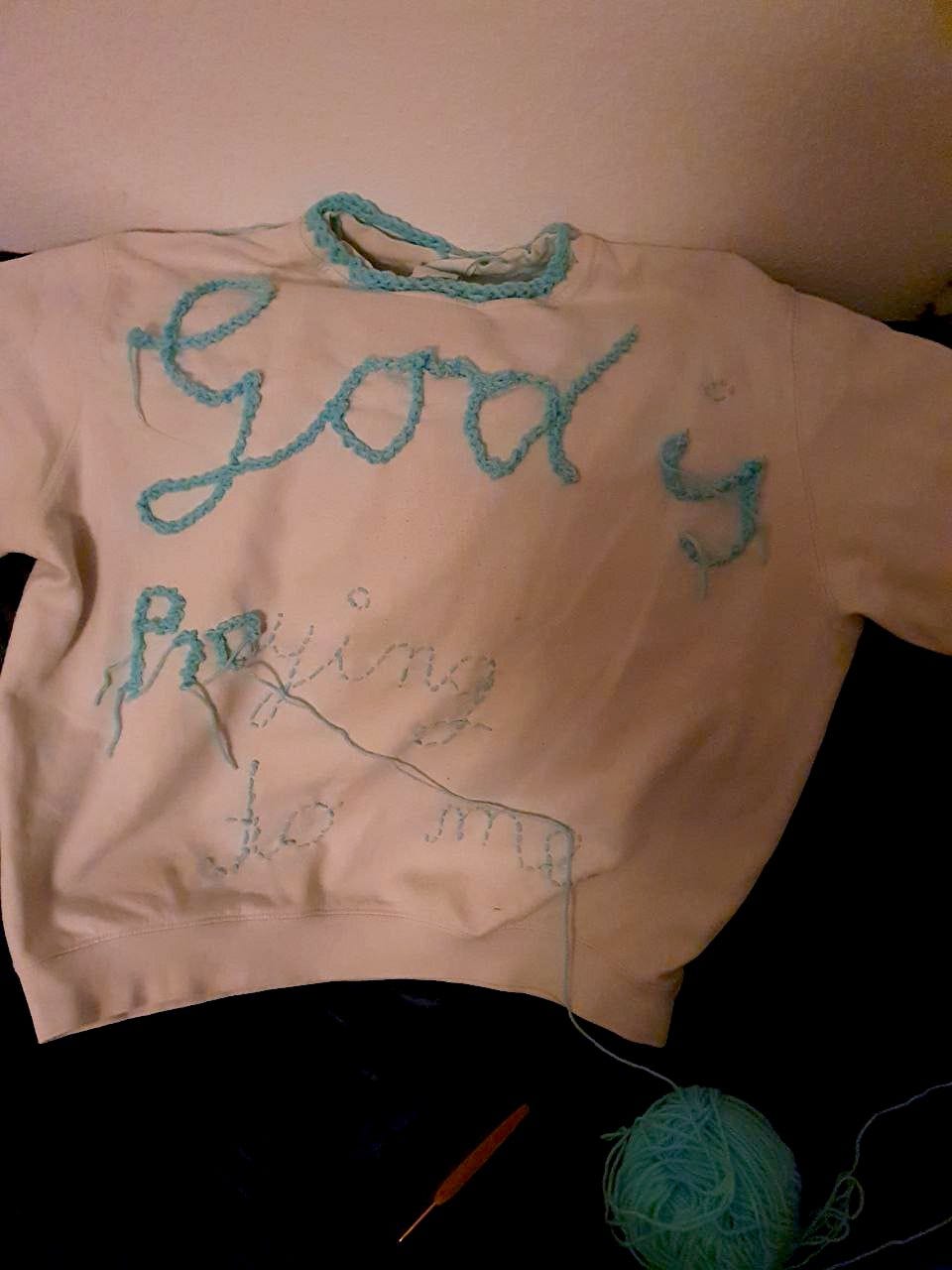 a white sweater is decorated with blue crocheting and stitching. the writing says God is praying to me