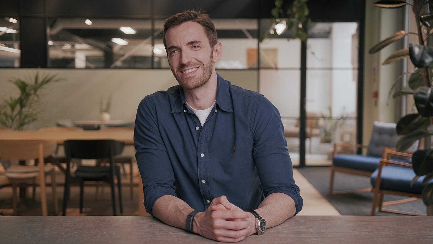 This is a photograph of Ian Wharton. Ian is a white man with short brown hair and beard. He is wearing a teal blue shirt, resting his hands on a table bench, and smiling as he looks slightly past the camera. Behind Ian is what appears to be an empty but stylish office space with chairs, a large table, and plenty of indoor plants.