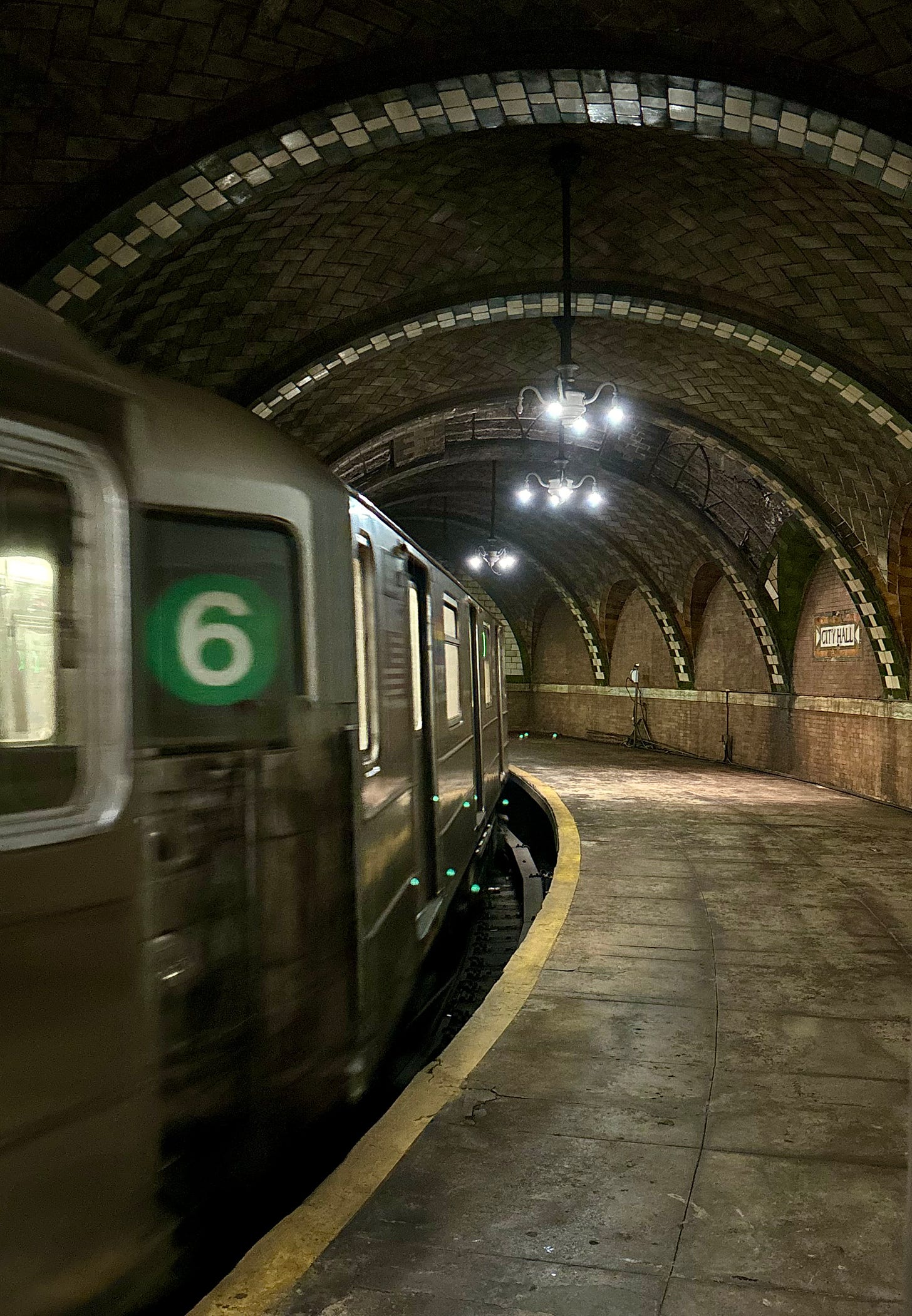 A 6 Train passes through a station with tiled arches and chandeliers hanging from the ceiling. On the wall is the name City Hall.