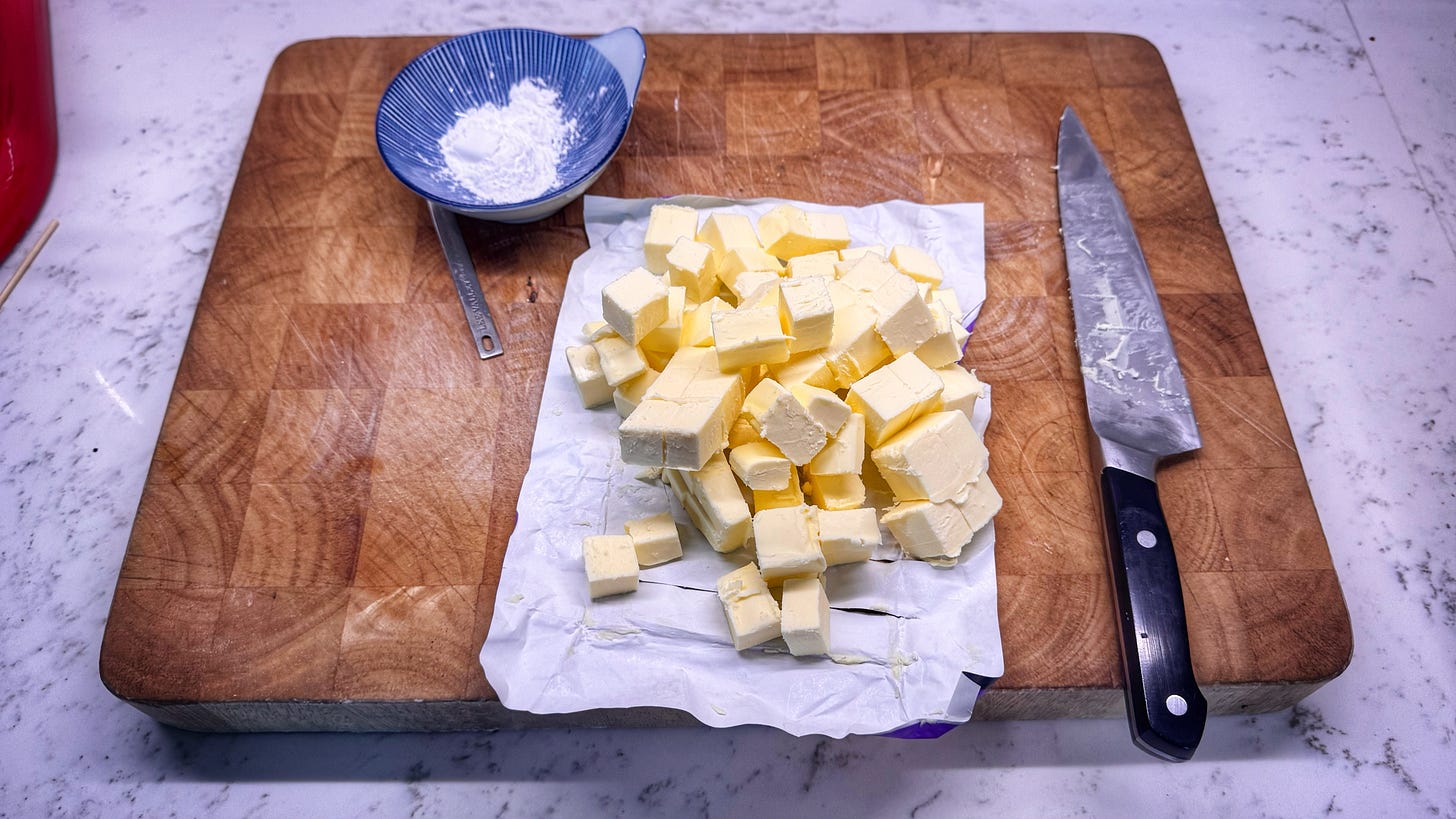 On a cutting board, a small bowl holding cornstarch, a pound of butter cut into small cubes, a chef's knife covered in butter residue