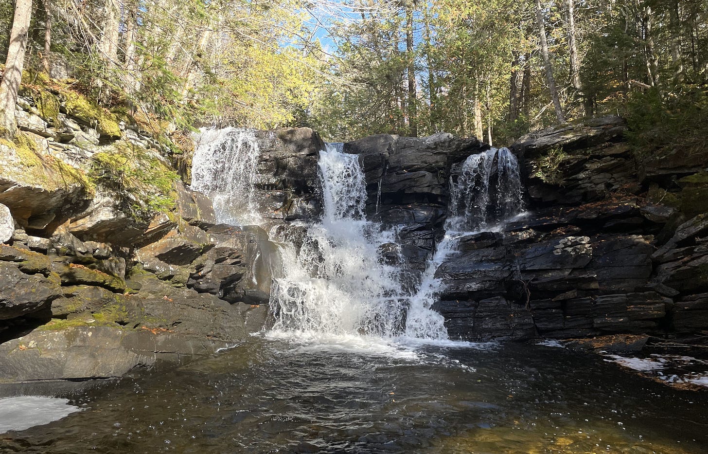 a three-part waterfall cascading through layered rock with sparse fall trees above