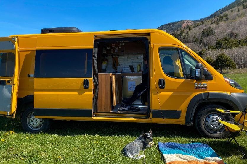 Hermes the yellow camper van sits in front of a small mountain with green grass underneath and blue sky above. The siding slide door is open, and Scout the blue heeler lounges in front of it