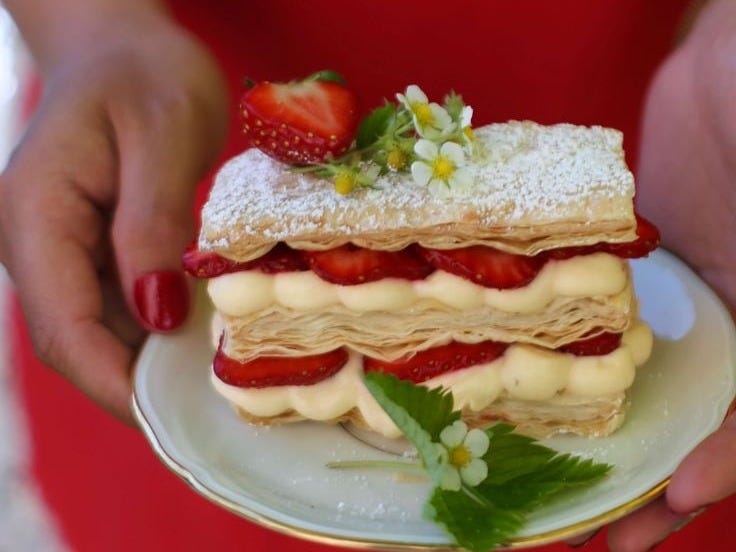A person in a red dress with painted red nails holds a delicate mille-feuille pastry layered with strawberries, cream, and puff pastry, topped with powdered sugar, a halved strawberry, and small white edible flowers, served on a white plate with gold trim.