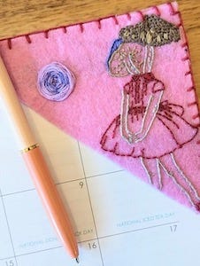 a pink embroidered bookmark positioned on the corner of a desk calendar. The pattern is of a young girl in a dress with a mushroom for a head