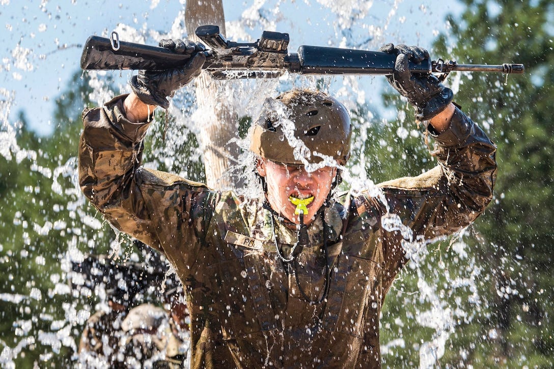 A cadet wearing protective gear raises a rifle overhead as water pours down.