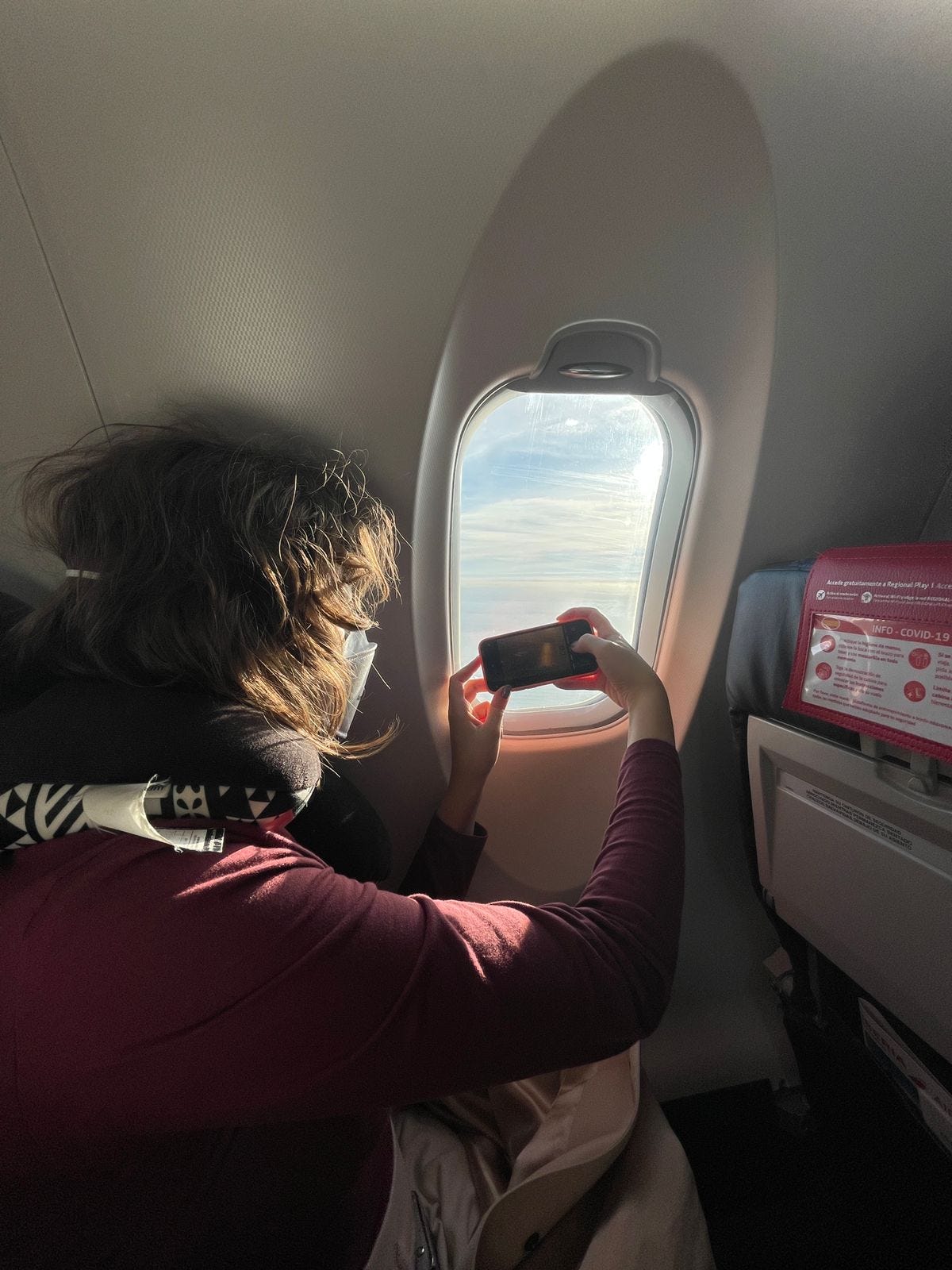 Foto em que estou dentro do avião totalmente descabelada, encolhida na janela tentando tirar uma foto do céu com o celular.