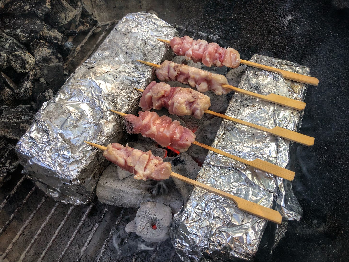 Skewers of chicken cooking over charcoal. The skewers are balanced between two bricks wrapped in aluminum foil. Everything is set in the bottom of a kettle grill. 