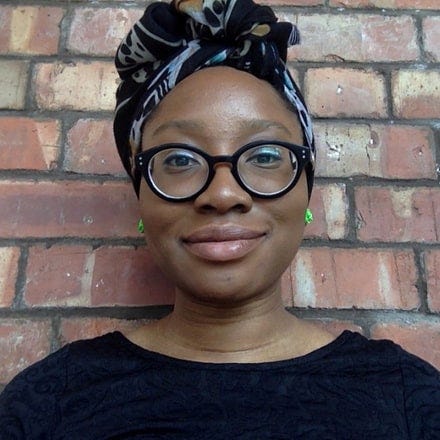 A Black woman faces the camera against a red brick background. She is wearing a black patterned head wrap, thick rimmed black circular glasses and a black top.