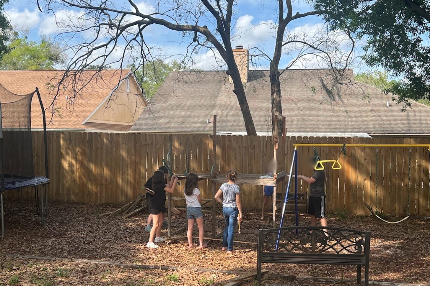  This image shows a group of children in a backyard engaged in what appears to be a group activity. They are surrounded by leafy trees, with residential houses in the background. One child in a dark top and shorts stands on a blue stool or ladder, seemingly reaching up towards something not visible in the picture, while another in a black shirt is close by, looking towards the first child. Three other children are to the right, with two of them looking at a piece of wood one of them is holding. Leaves and wood debris cover the ground, and there are elements of a playground, like swings, visible.