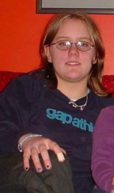Hannah, aged 14, has shoulder length blonde hair, glasses with thin black frames, a navy Gap Athletic hoodie with a stylised teal logo, black jeans, chipped nail polish and a plaster on her index finger. Her siblings are out of shot due to photo cropping. 