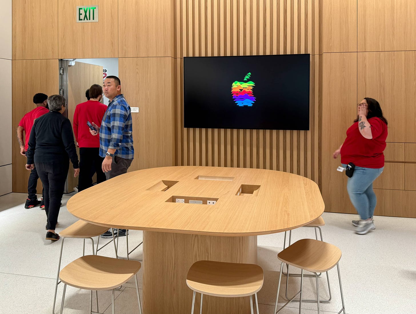 A Today at Apple table inside Apple South Shore. A large LED display is mounted on the rear wall of the store. The table is circled by stools.